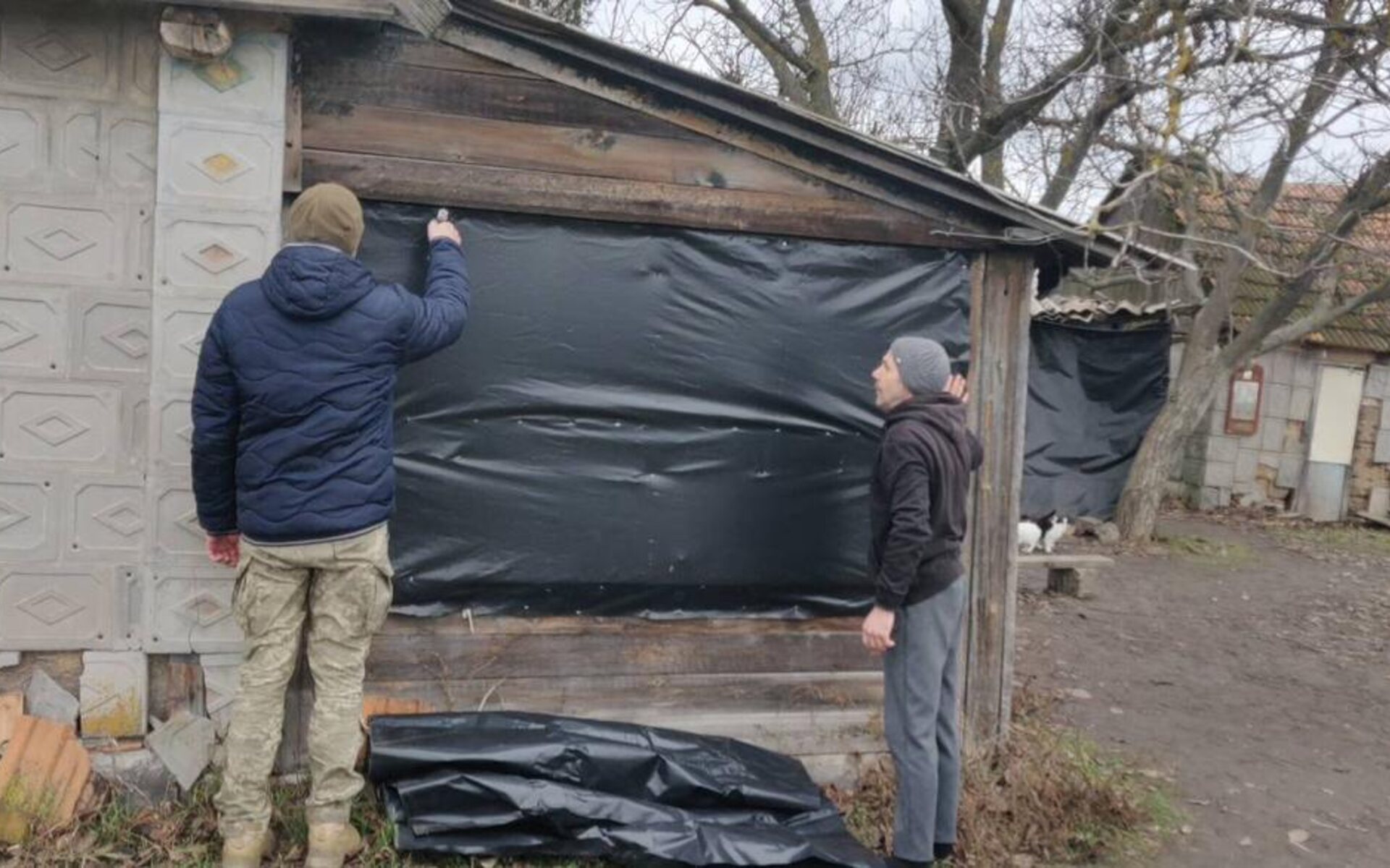 A soldier in camouflage and a civilian work side by side, sealing a window with sheeting on a damaged village house.