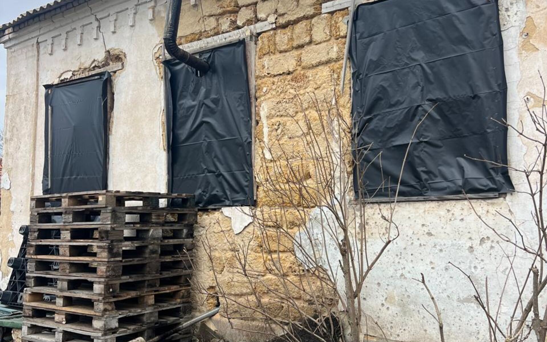 An old village house with cracked walls, windows sealed in black sheeting, and a freshly installed steel chimney.