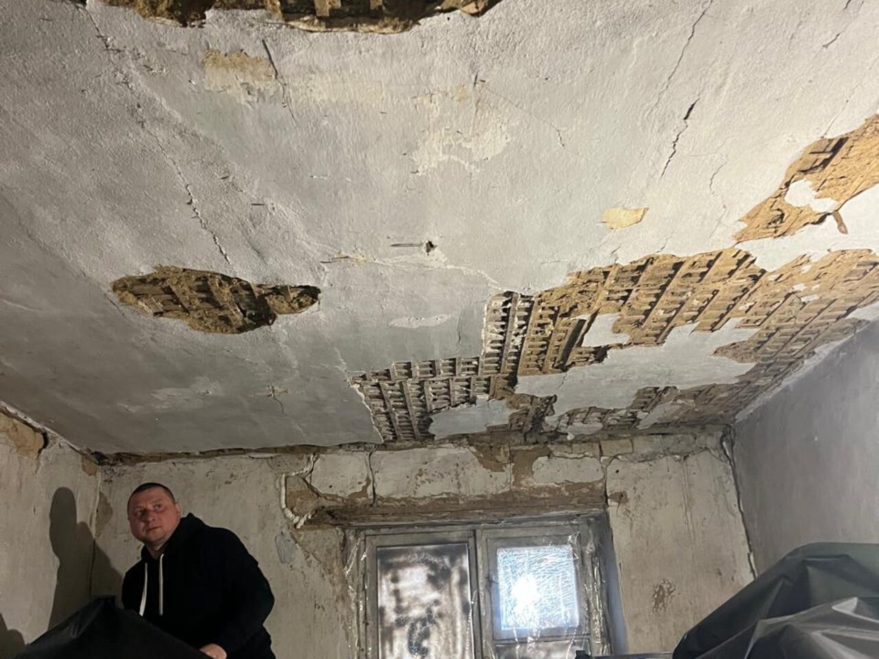A man inside a damaged house surveys a collapsed plaster ceiling with exposed wooden lath.