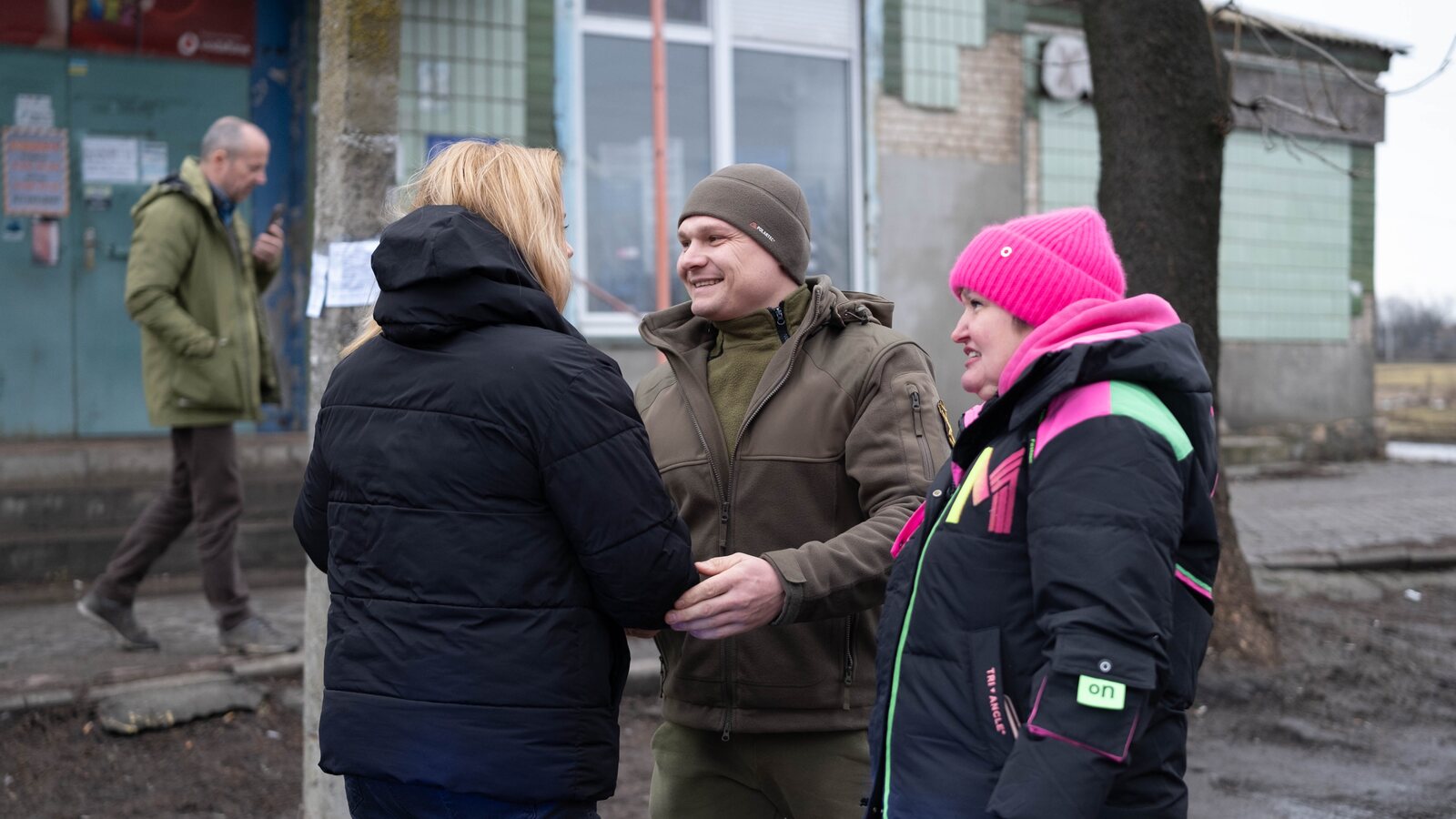 A soldier in fleece smiles with two civilians in winter coats during a handover visit on a village street.